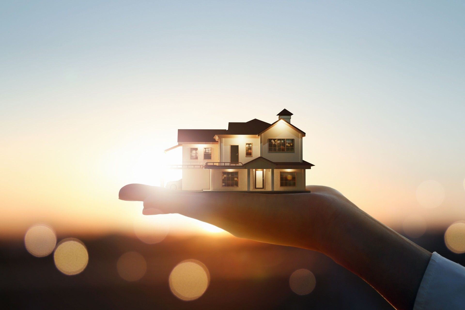 Hand holding a miniature house against a sunset background, symbolizing real estate and home ownership.