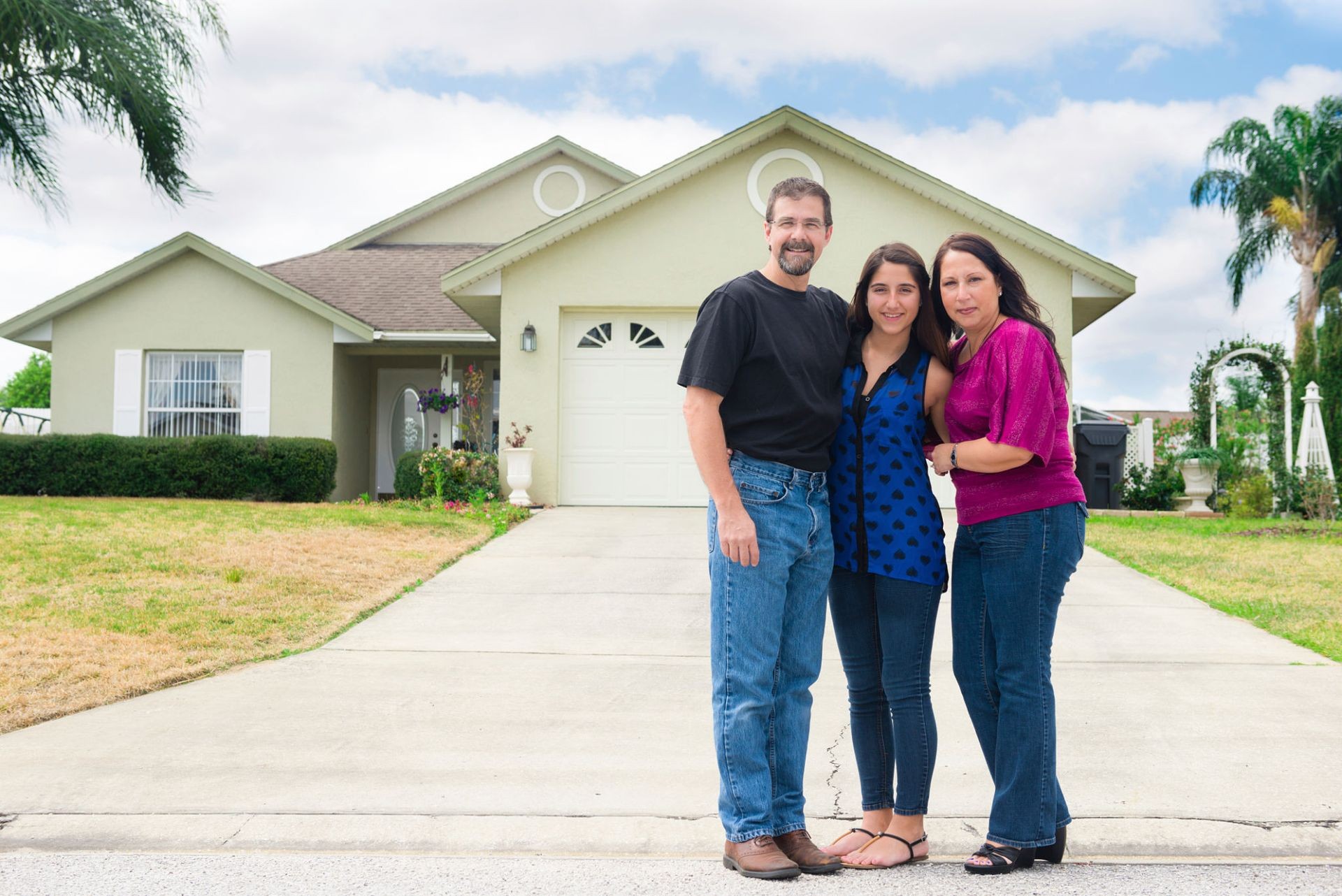 Family of three smiling in front of their house with a green lawn and driveway.