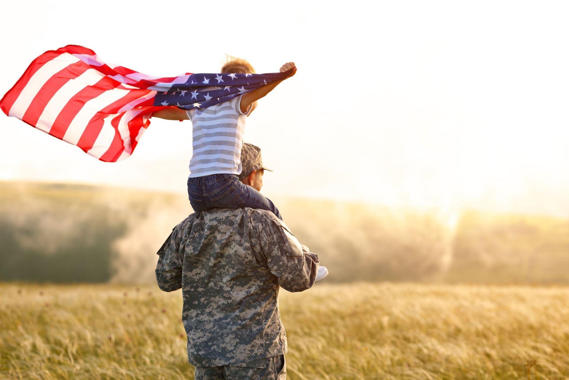 A child sits on a soldier's shoulders, holding an American flag in a field during sunset.