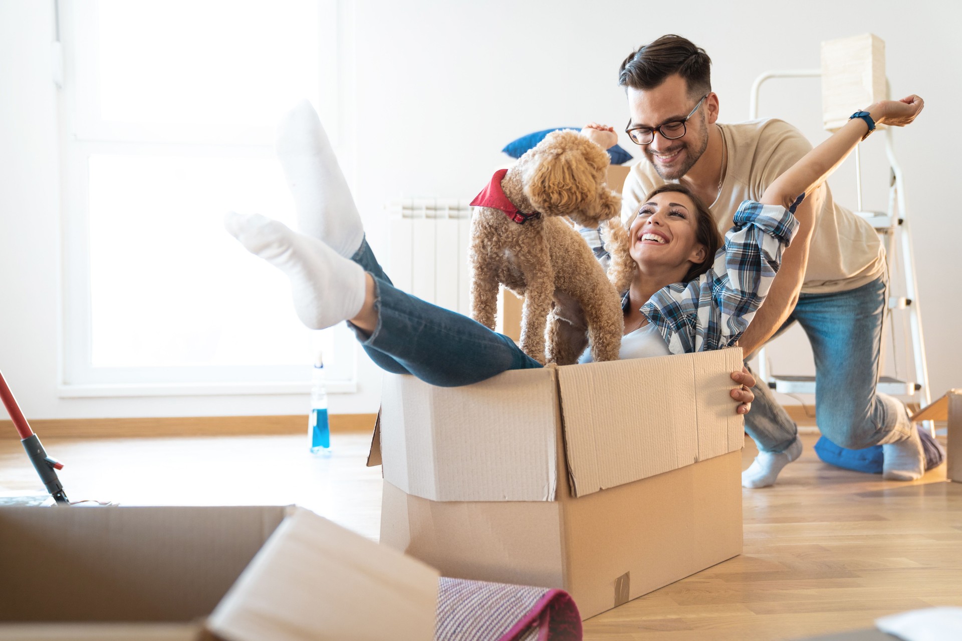 Cheerful young couple being silly having fun while unpacking boxes at new home, together with their dog